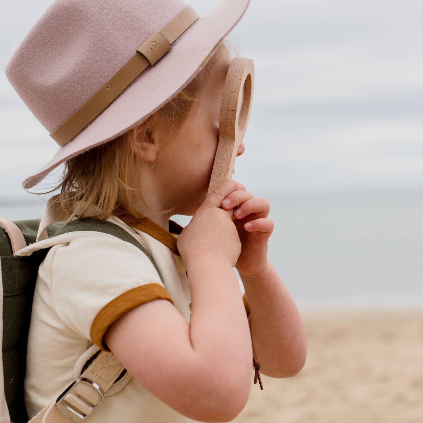 Child wearing a pink hat and beige outfit with a backpack, standing on a beach, looking through a magnifying glass