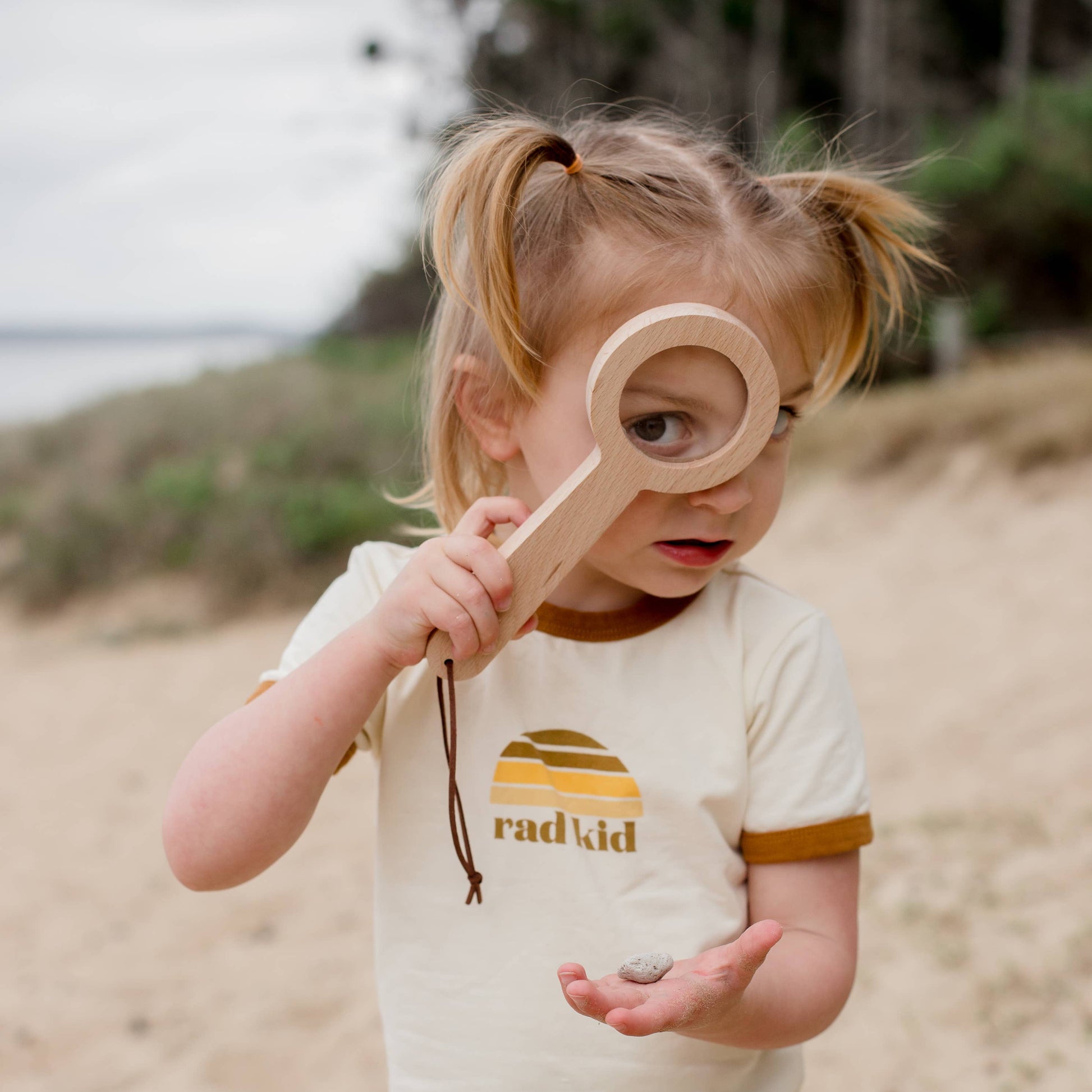 Child on a beach holding a wooden magnifying glass toy, wearing a 'rad kid' t-shirt.