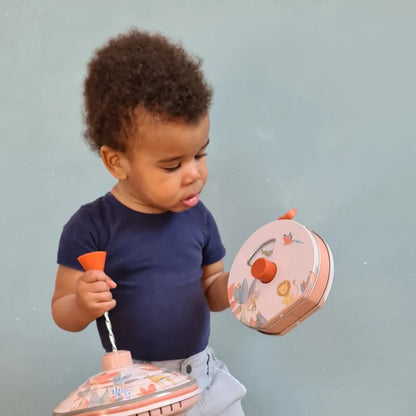 A toddler kneeling on a wooden floor against a light blue-gray wall, holding a pink and orange toy drum and a matching toy cup with a decorative floral pattern.