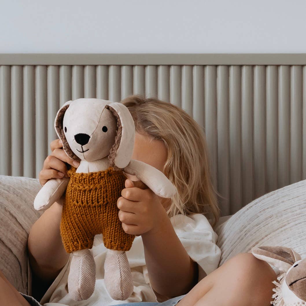 Child holding a plush dog toy with knitted overalls in a cozy indoor setting.