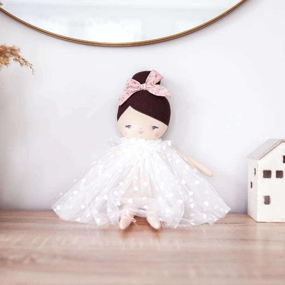 Stuffed doll with a white dress and pink bow on a wooden surface with a white wall background