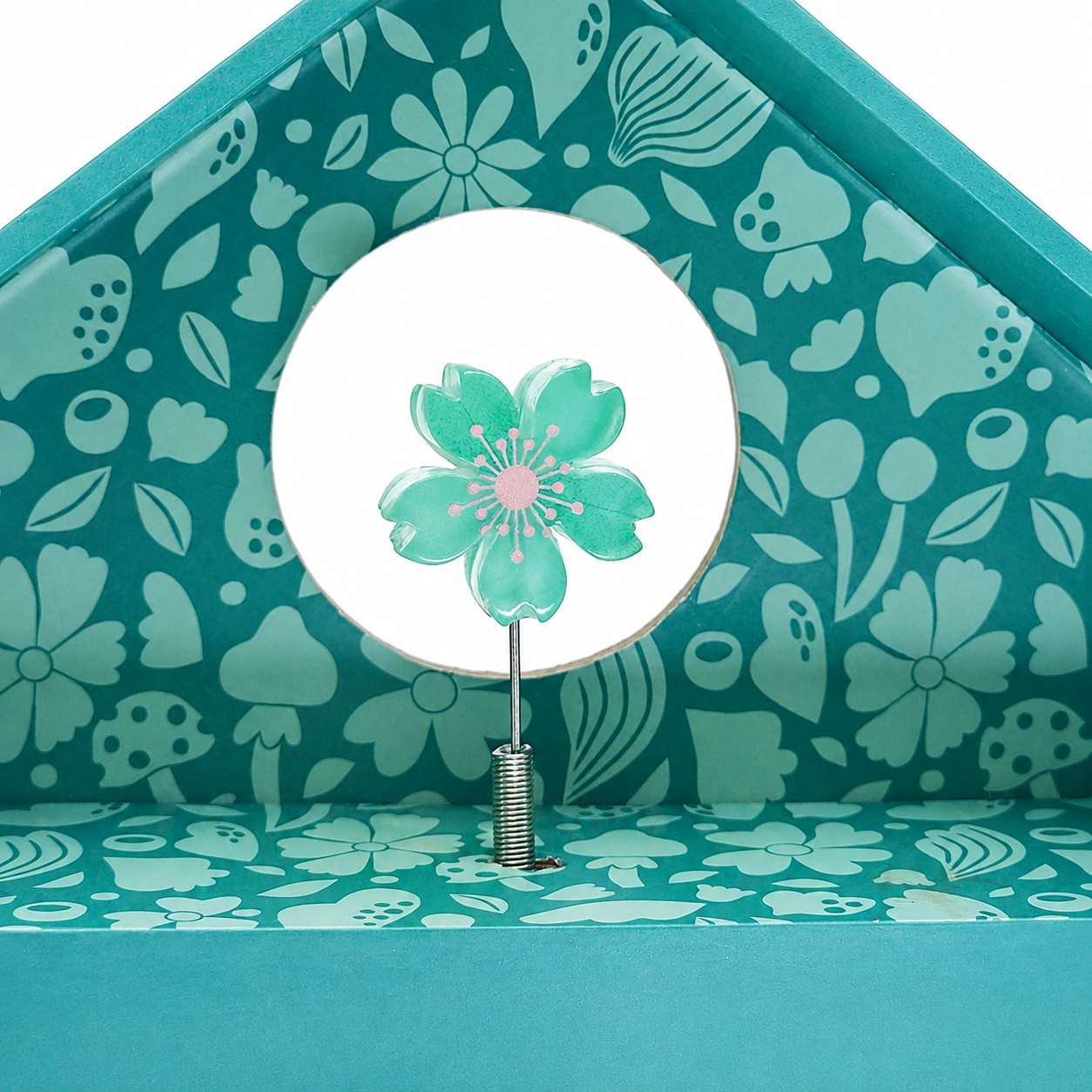 A decorative teal and mint floral house-shaped box with a small flower-shaped flower in the center, featuring a white circular insert and a patterned interior.