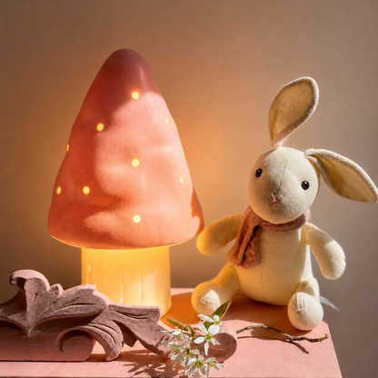 A warm-toned mushroom-shaped night light with glowing cutout lights, styled beside a cream plush bunny with a pink scarf on a pink surface against a warm beige background.