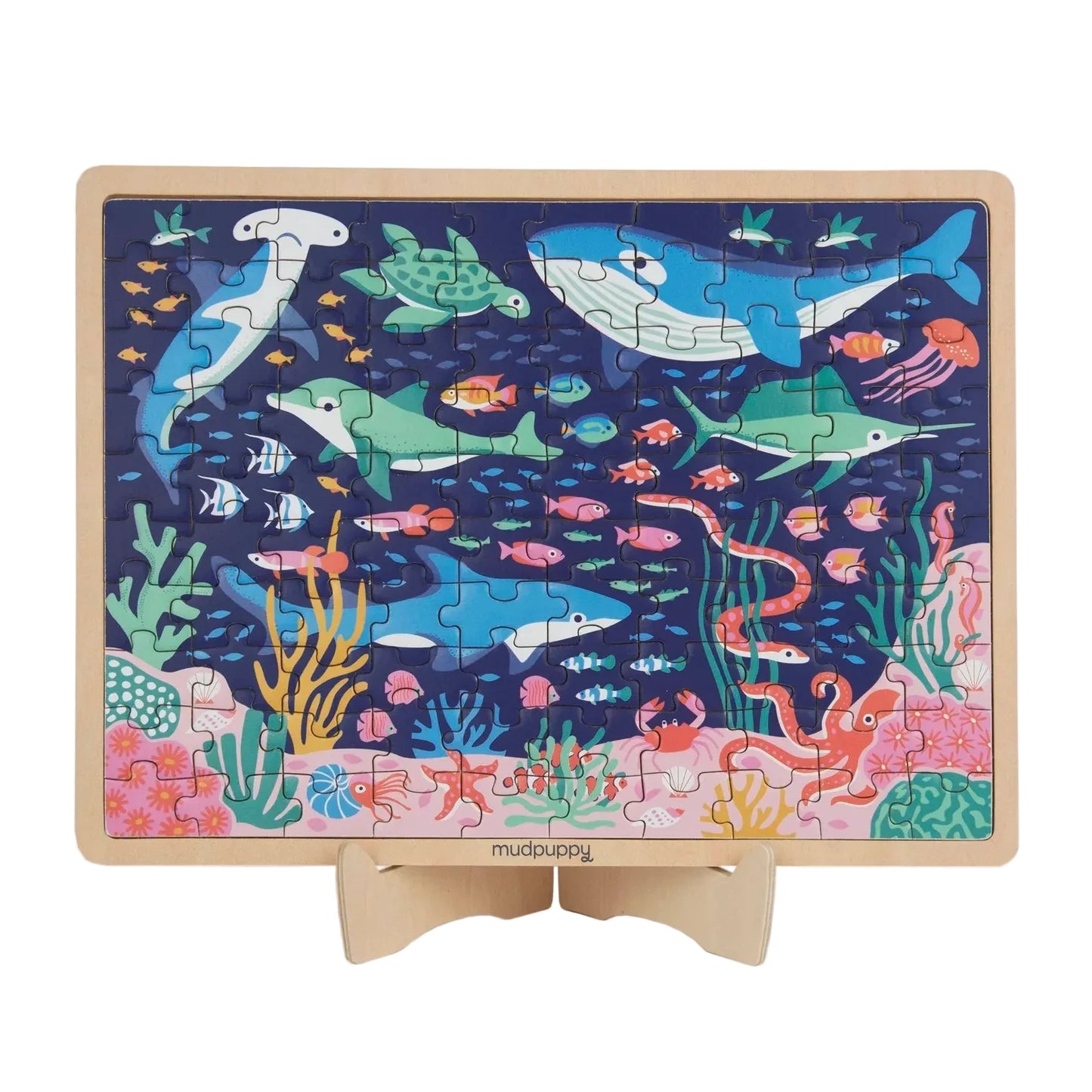 A rectangular wooden children's jigsaw puzzle on a small wooden stand, featuring a colorful underwater scene with sharks, dolphins, fish, coral, and sea life on a dark blue background.