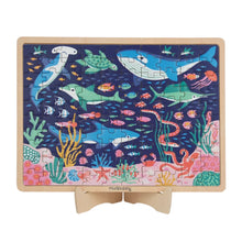 A rectangular wooden children's jigsaw puzzle on a small wooden stand, featuring a colorful underwater scene with sharks, dolphins, fish, coral, and sea life on a dark blue background.