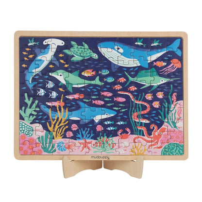 A rectangular wooden children's jigsaw puzzle on a small wooden stand, featuring a colorful underwater scene with sharks, dolphins, fish, coral, and sea life on a dark blue background.