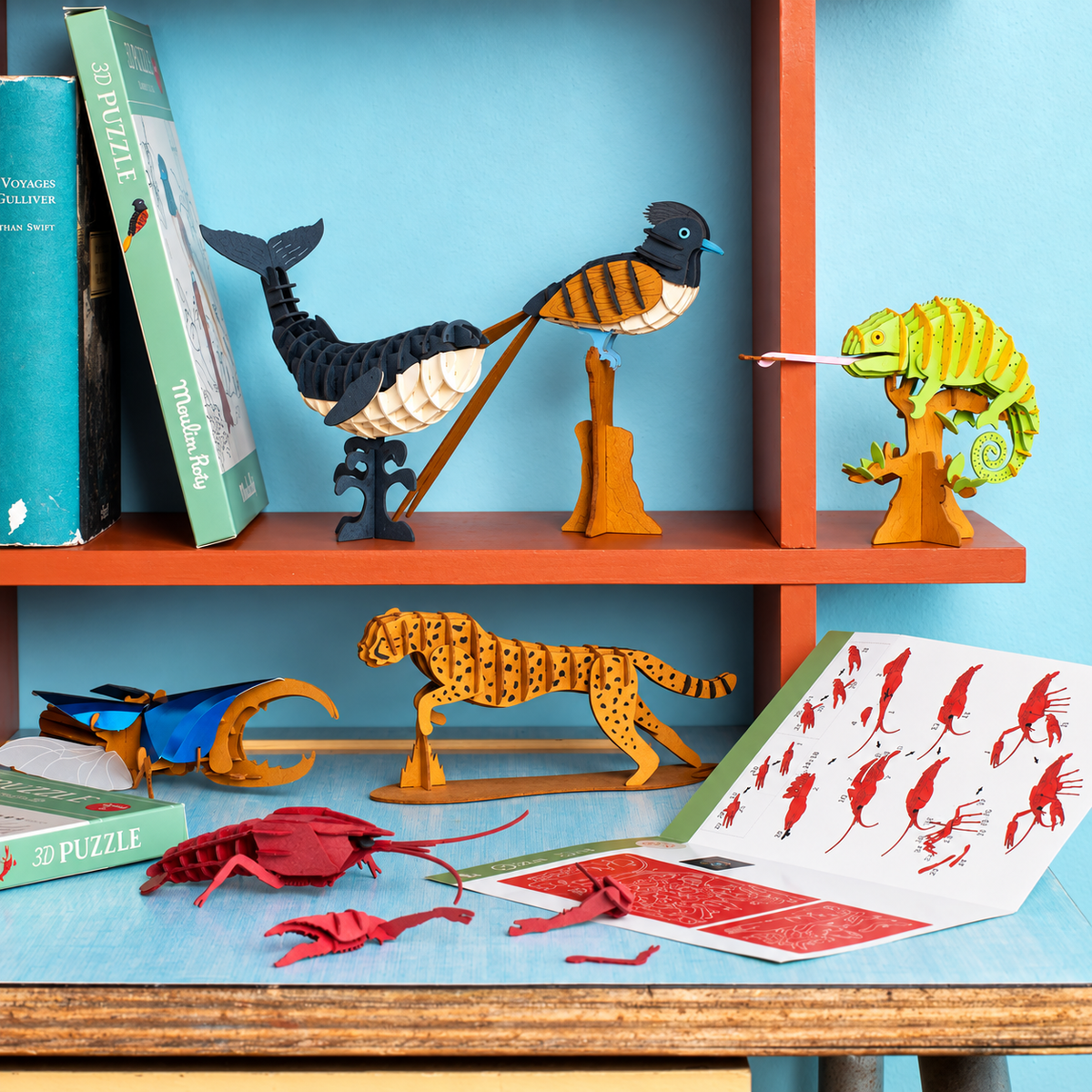 A styled tabletop display of wooden animal puzzle boxes and assembled wooden animal figures, including a bird, chameleon, beetle, cheetah, and red lobster, arranged on a shelf and table against a light blue wall.