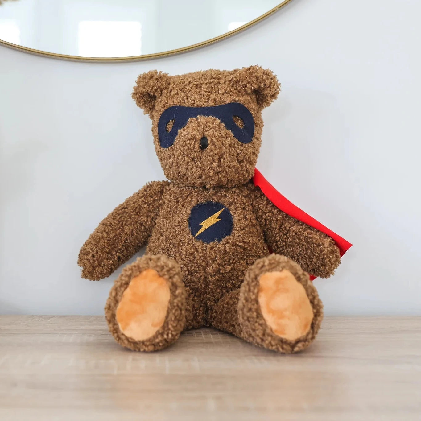 Brown teddy bear with superhero cape and mask on a wooden surface.