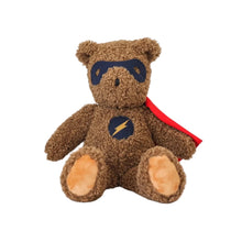 Brown teddy bear with superhero cape and mask on a wooden surface.