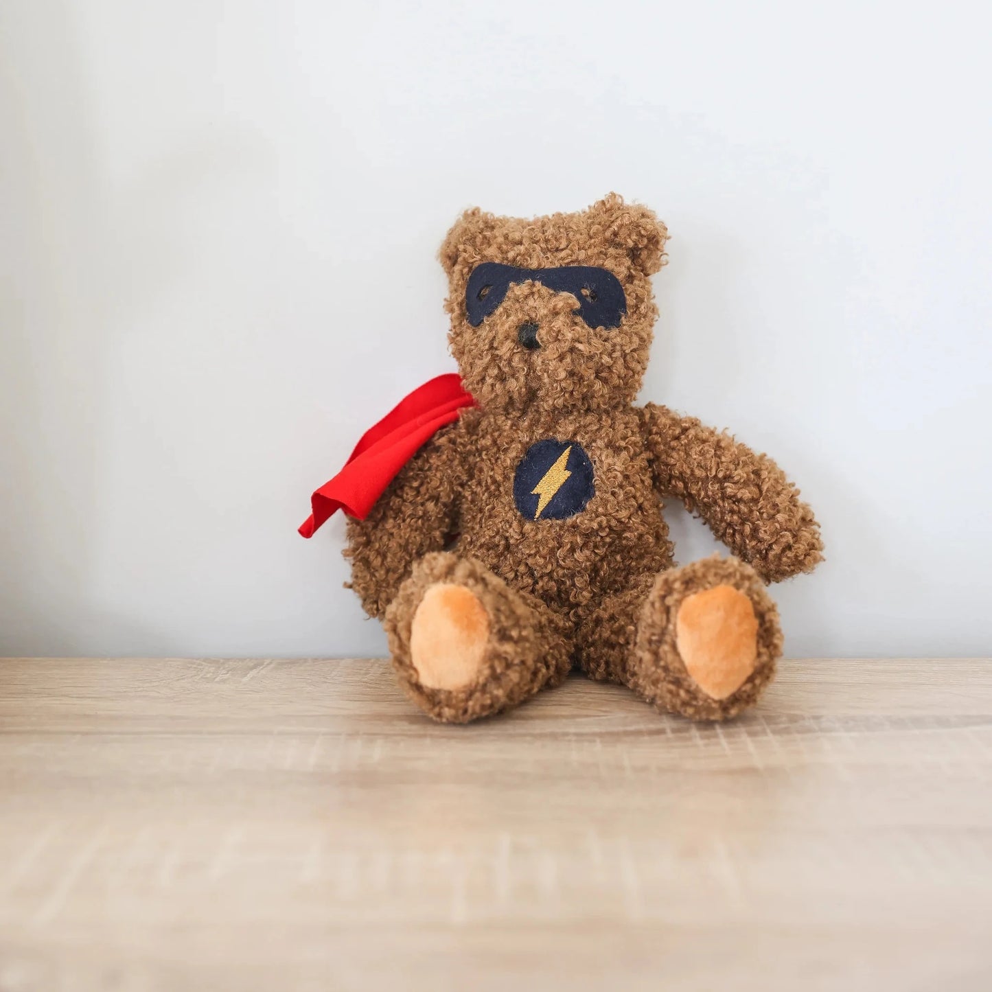 Teddy bear with a superhero cape on a wooden surface with a white wall background