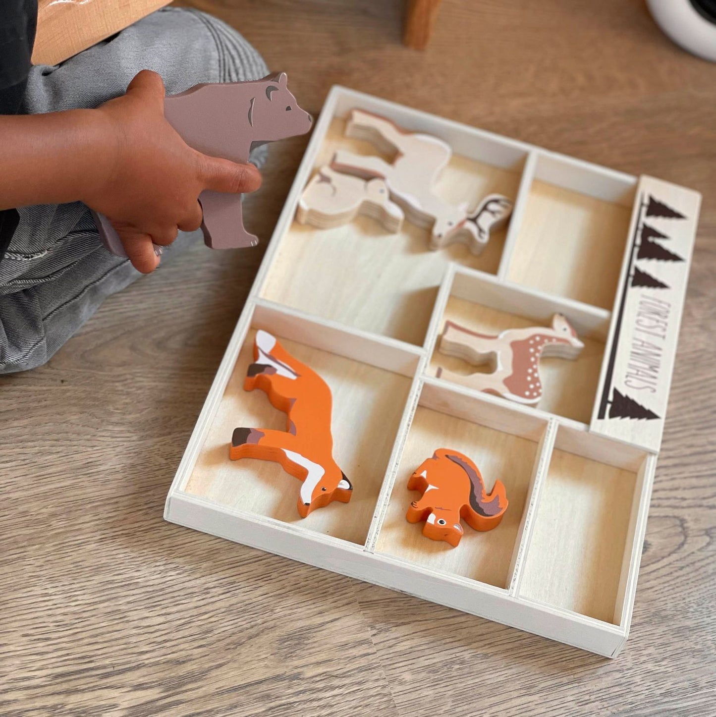 Child playing with wooden animal toys in a box on a wooden floor.