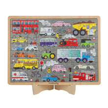 A children's wooden traffic-themed jigsaw puzzle displayed on a small wooden stand. The puzzle features a gray background with various colorful vehicles including a red double-decker bus, yellow dump truck, school bus, ambulance, fire truck, train, and cars, with the brand name 'mudpaisley' printed at the bottom.
