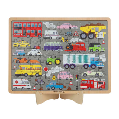 A children's wooden traffic-themed jigsaw puzzle displayed on a small wooden stand. The puzzle features a gray background with various colorful vehicles including a red double-decker bus, yellow dump truck, school bus, ambulance, fire truck, train, and cars, with the brand name 'mudpaisley' printed at the bottom.
