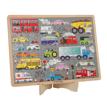 A children's wooden traffic-themed jigsaw puzzle displayed on a small wooden stand. The puzzle features a gray background with various colorful vehicles including a red double-decker bus, yellow dump truck, ambulance, school bus, fire truck, and other cars and trucks, with the brand name 'mudpuppy' visible on the front of the stand.