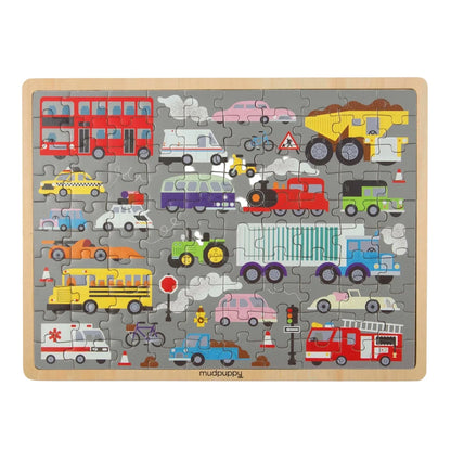 A children's wooden jigsaw puzzle featuring colorful cartoon vehicles on a gray background, including buses, cars, trucks, a train, a fire truck, an ambulance, and a bicycle. The puzzle is assembled and photographed from above on a clean white background.