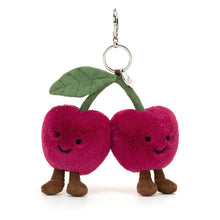 A plush toy resembling two cherries with faces, legs, and a green leaf on a silver clasp.