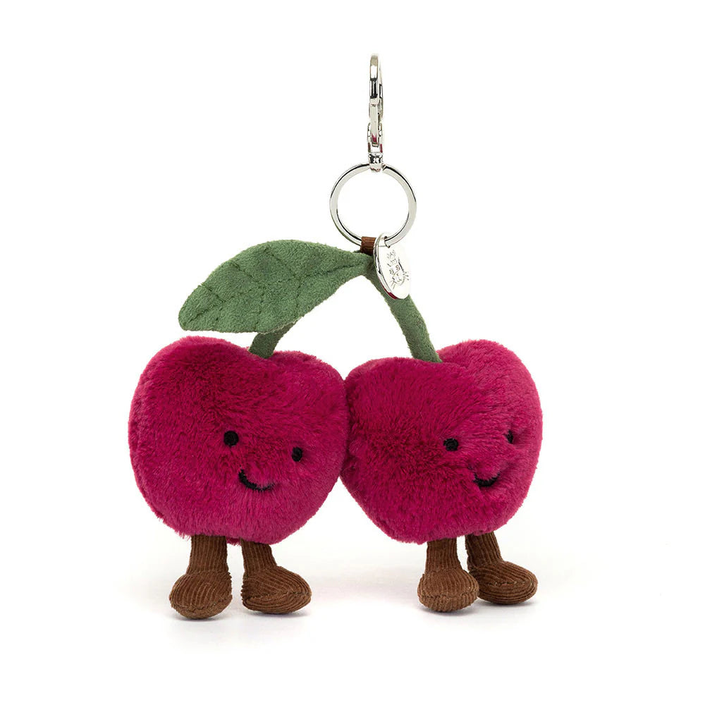 A plush toy resembling two cherries with faces, legs, and a green leaf on a silver clasp.