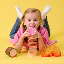 A child lying on a yellow background with various colorful coil spring toys, with one toy resembling a pizza slice, another like a donut, and a third in a bright orange color.