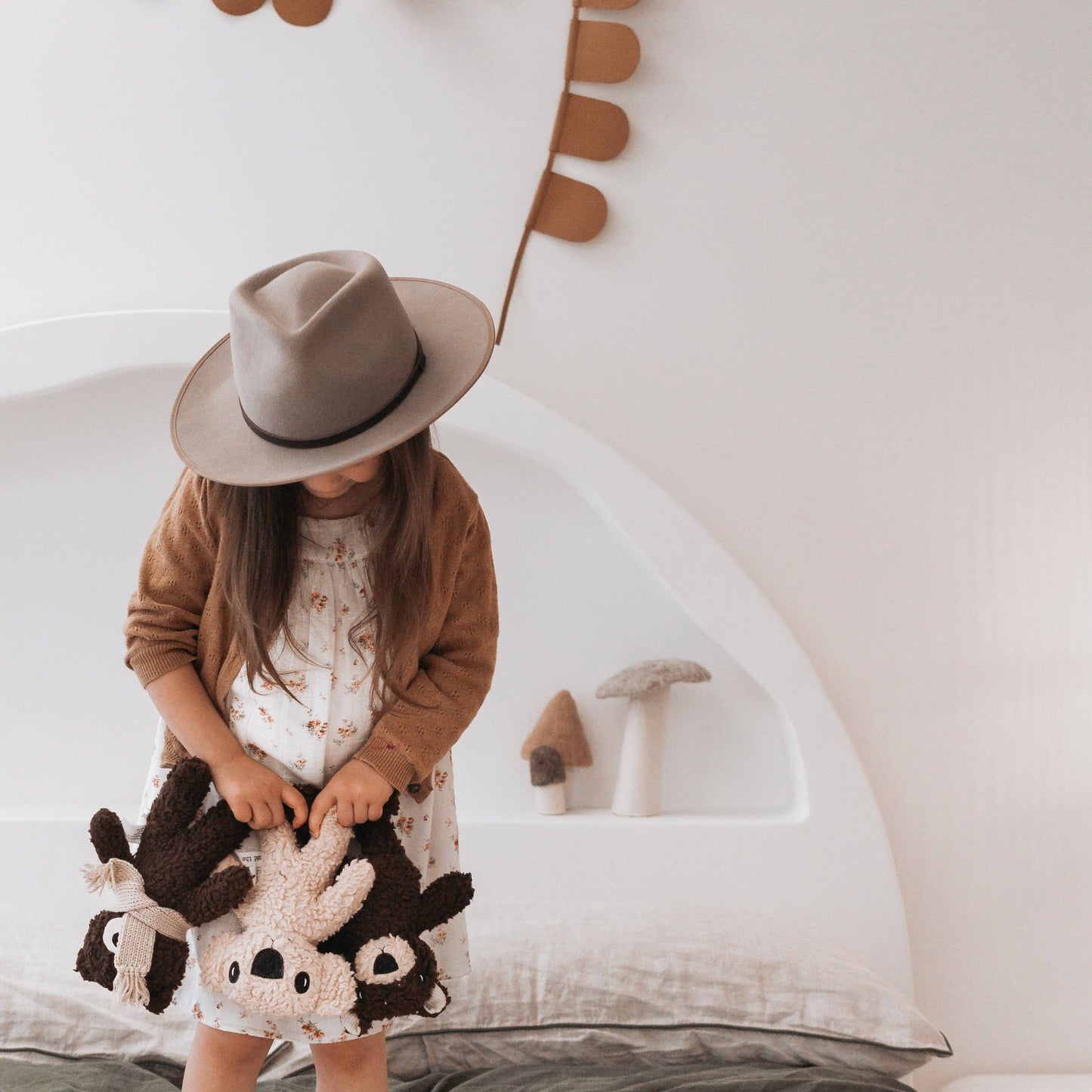 Child wearing a hat and holding plush toys in a cozy indoor setting
