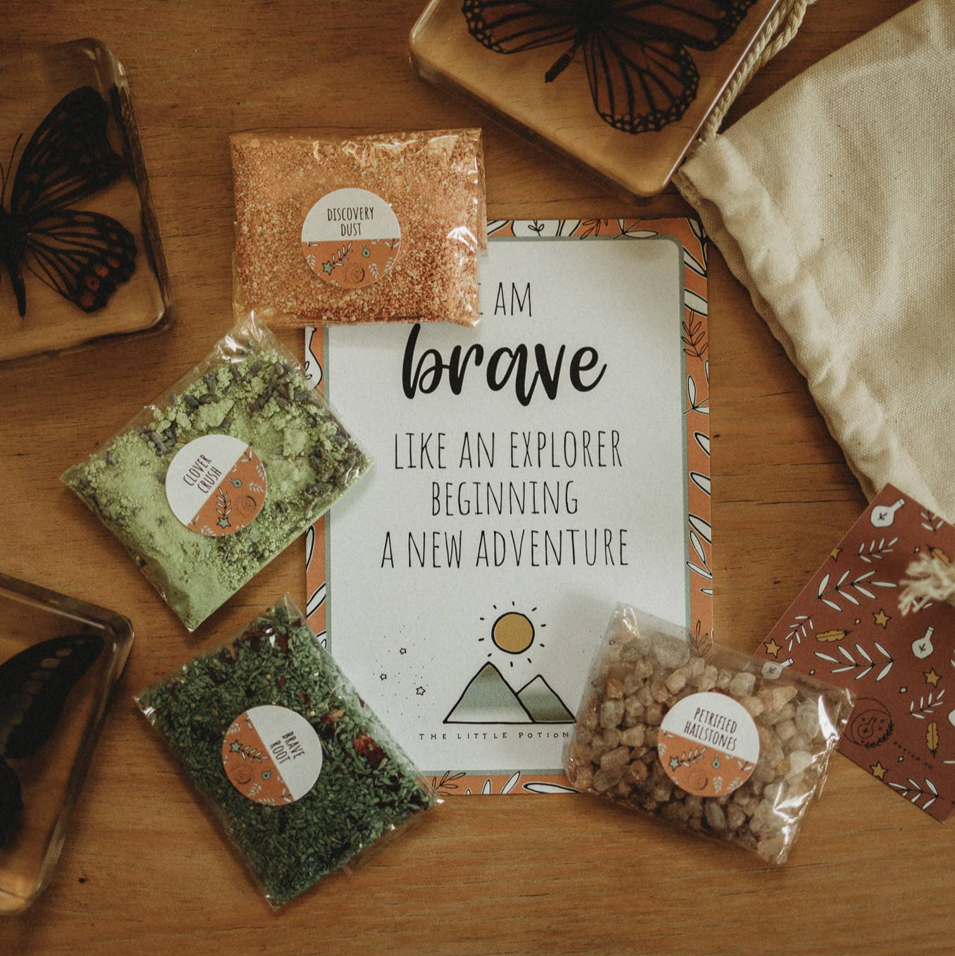 The image displays the Heart of an adventurer - Potion Pouch product, which includes four sparkly dry ingredients in biodegradable cellophane bags, a potion card, and a linen cloth bag, all laid out on a wooden surface with a magical theme.