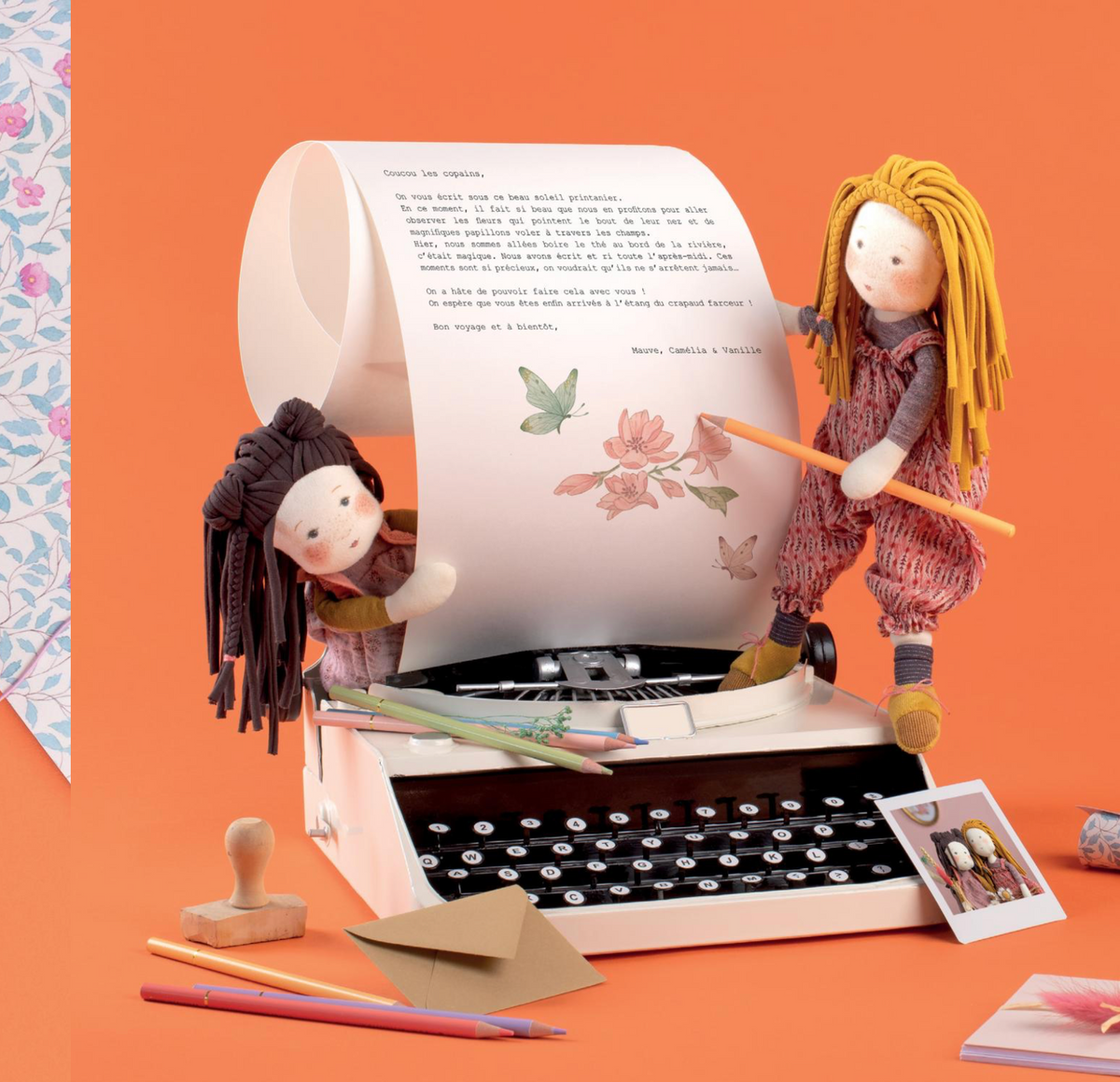A styled tabletop scene with two handmade doll figures around a vintage typewriter that is printing a letter. The setting includes pens, an envelope, a rubber stamp, a small photo card, and patterned paper on a solid orange background.