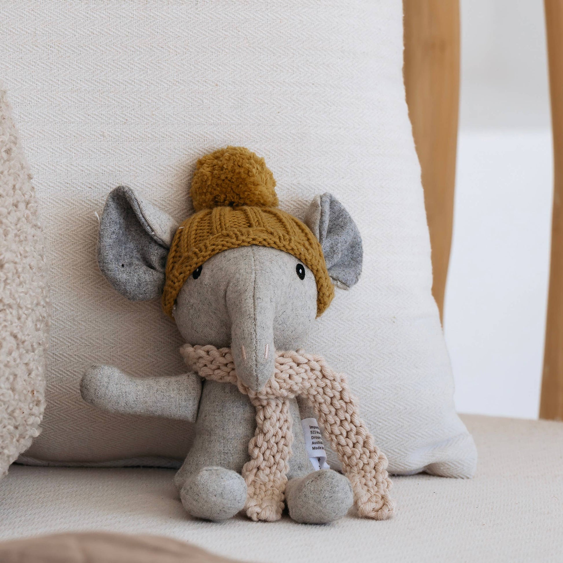 Stuffed elephant toy wearing a yellow knitted hat and beige scarf on a white pillow.