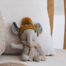 Stuffed elephant toy wearing a yellow knitted hat and beige scarf on a white pillow.