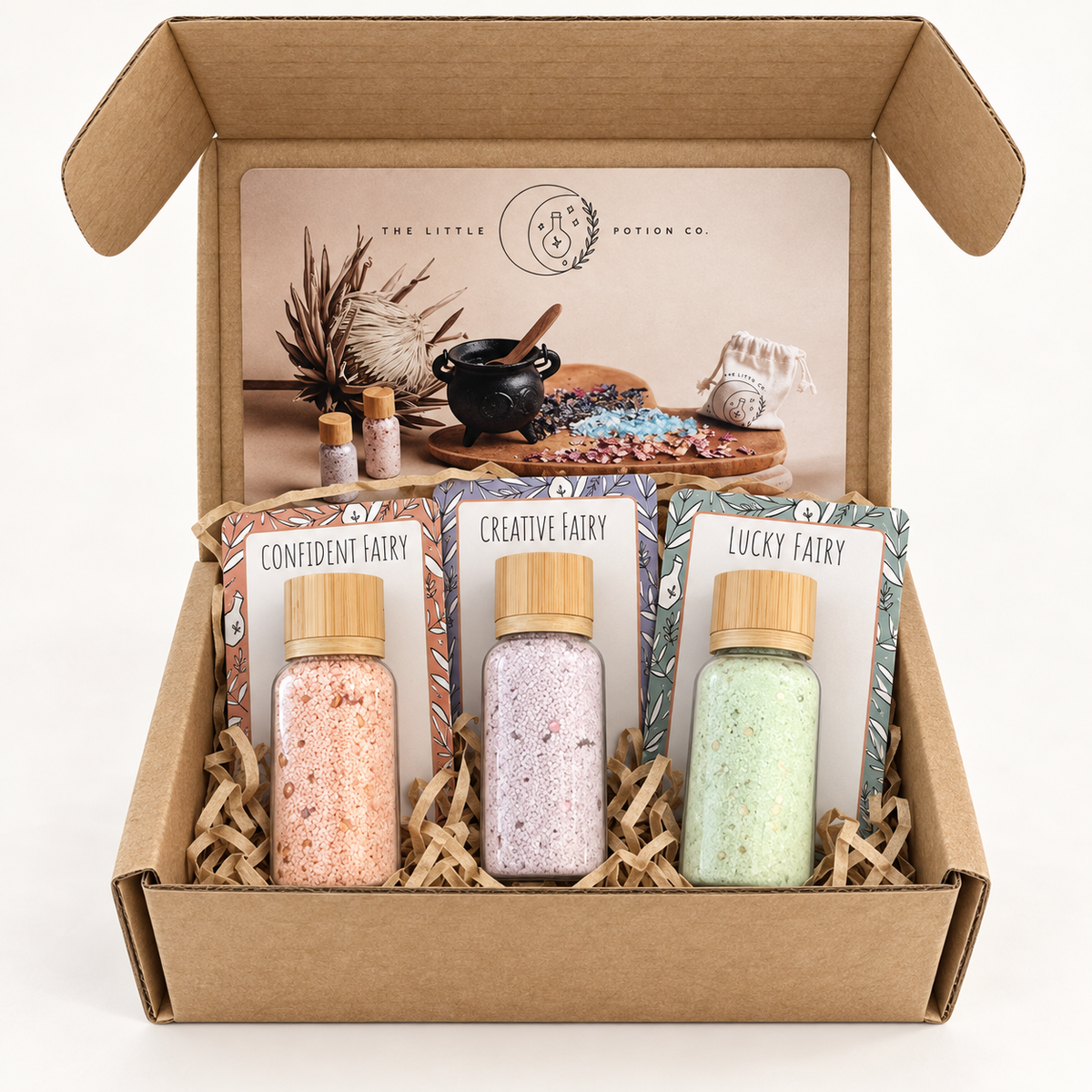 An open kraft gift box from The Little Potion Co. containing three small glass bottles of bath salts in pink, lavender, and mint green, each with a wooden cap and a decorative card labeled with a fairy name: Confident Fairy, Creative Fairy, and Lucky Fairy.