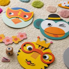 Assorted colorful cardboard animal face pieces for a matching game laid out on a beige surface, including pieces that resemble a fish, a monkey, and a chicken, along with various shaped pieces for eyes, ears, and beaks.
