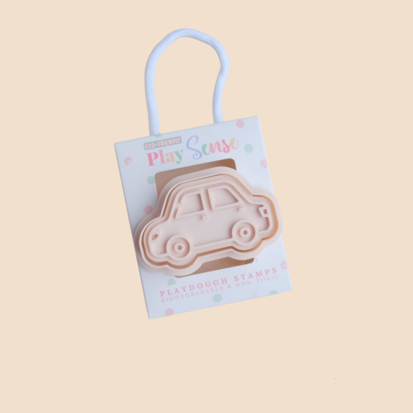A beige-colored playdough stamp shaped like a car, packaged in a box with a clear window displaying the product. The packaging is accompanied by a small wooden bead chain and a pink item in the background.