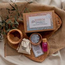 A mini potion kit laid out on a wooden surface, including a box labeled Rainbow Sparkles, dry fizz ingredients in a cotton pouch, sparkly liquid in a plastic bottle, a glass jar of refill liquid powder, and a mini affirmation and spell card.