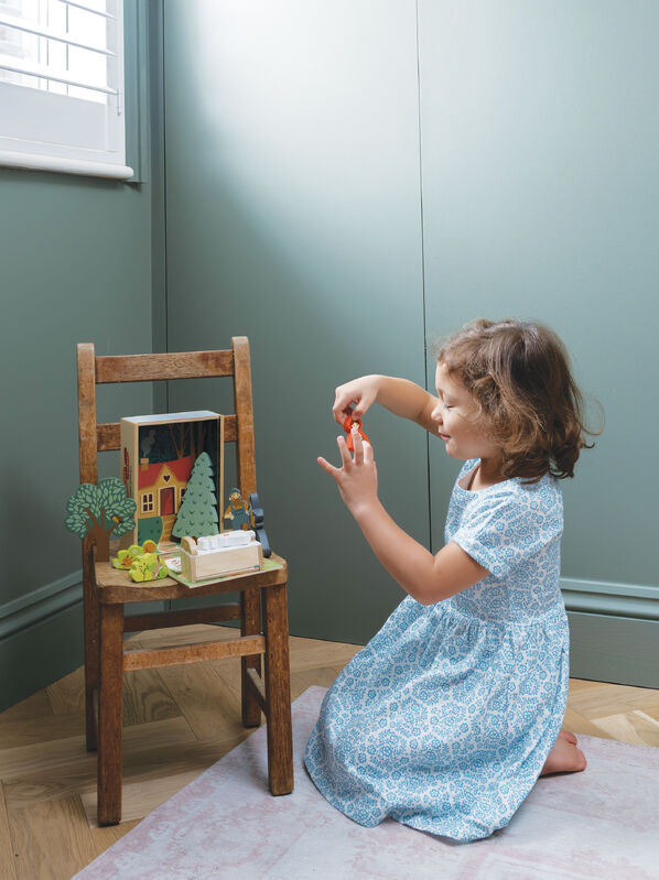 Child in a blue dress playing with toys on a wooden chair against a green wall.