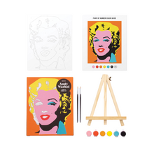 A paint by numbers kit featuring a silkscreen portrait of Marilyn Monroe by Andy Warhol, including a wooden easel, a paint-by-numbers artboard, two paintbrushes, and six small tubs of acrylic paints.