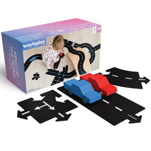 A child playing with a road track set that includes black track pieces and two wooden cars, one blue and one red, on a patterned play mat.