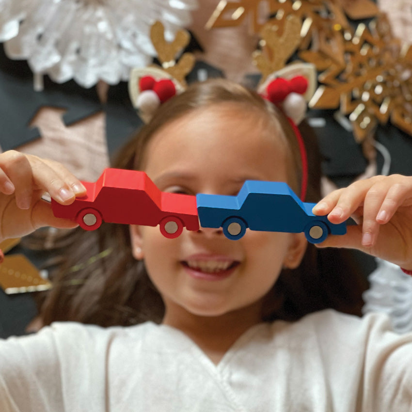 Child holding up two wooden vehicles, one red, one blue.