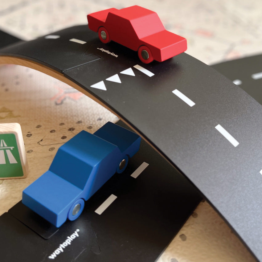 Red and blue wooden cars pictured on a black roadway track with white lines