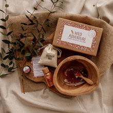 A Mini Wild Adventure Potion Kit displayed on a burlap fabric with a wooden bowl, containing dry fizz ingredients in a cotton pouch, sparkly liquids in plastic bottles, a biodegradable refill liquid powder bag, and a mini affirmation card.