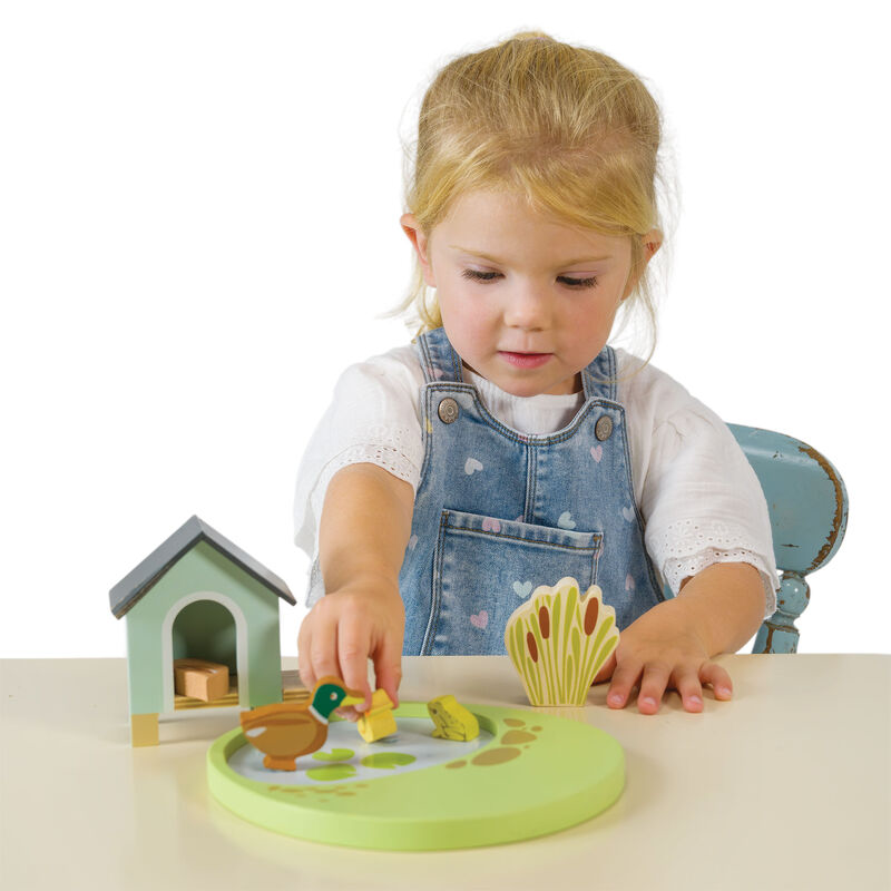 A young child wearing denim overalls and a white shirt sits at a table playing with a wooden toy set. The set includes a round green base with animal pieces, a small house, and a fan of animal-shaped pieces, photographed against a clean white background.
