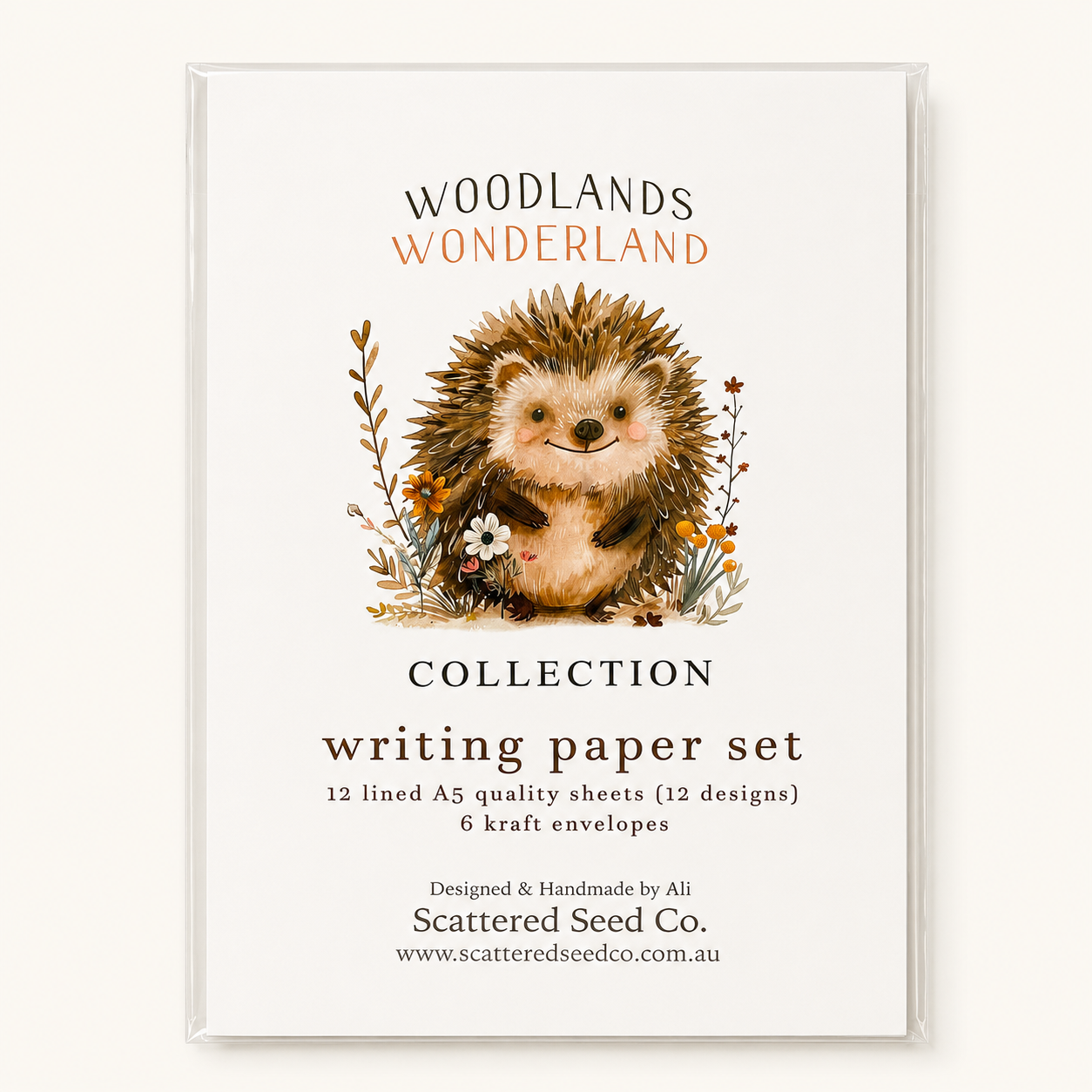 A clear acrylic sleeve encloses a printed card for a writing paper set. The card reads 'Woodlands Wonderland Collection writing paper set' with an illustration of a hedgehog and floral details, and shows the brand 'Scattered Seed Co.