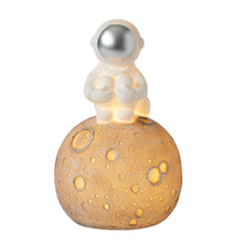 An astronaut-themed night light featuring a small figurine of an astronaut on a spherical base with a warm LED bulb, set against a moonlit background.