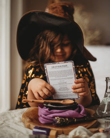 Child in witch costume holding a card with text, sitting at a table with a bottle and other items.