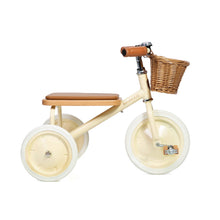 A cream-colored tricycle with a brown seat, oak wood pedals, and a front wicker basket, set against a white background.