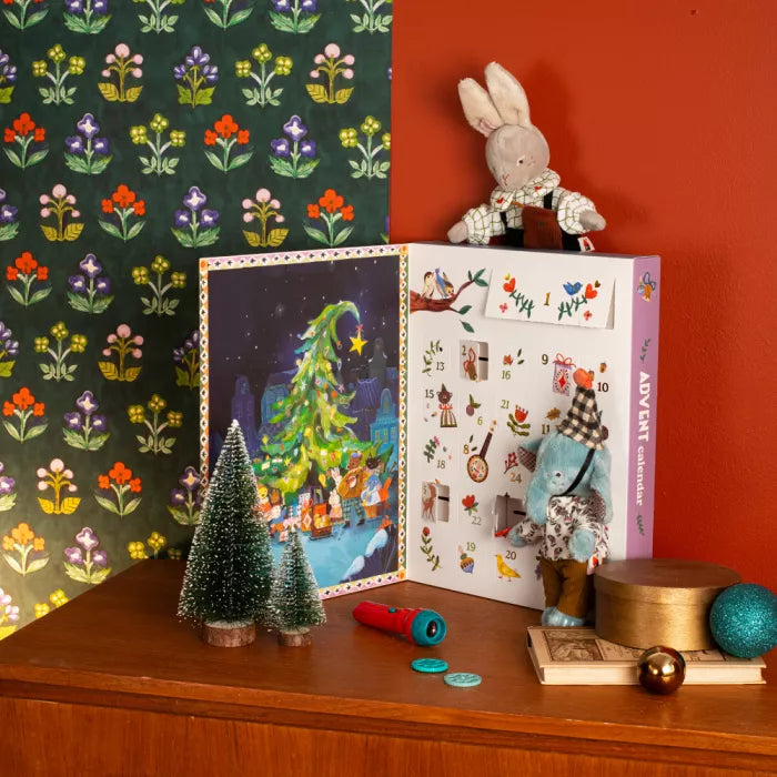 Decorative setup with a Christmas-themed child's advent calendar book, small trees, and a rabbit figurine on a wooden surface.