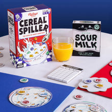 The image shows the Cereal Spiller Game, including the packaging of the game box, cereal bowls, and various game tokens placed on a playing board. A glass of orange juice is also visible, suggesting the cereal theme of the game.