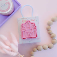 A pink castle-shaped playdough stamp on a packaging card, with a small purple container and decorative elements in the background.