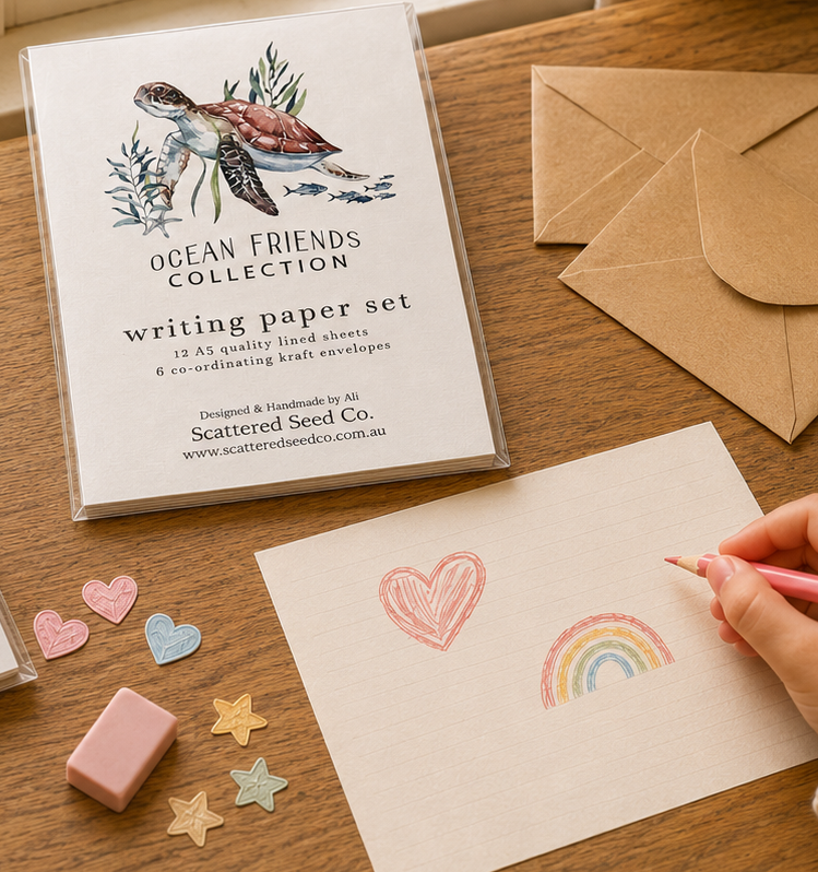 A hand uses a pink pencil to draw a heart and rainbow on white writing paper, with a packaged Ocean Friends Collection writing paper set in the background and kraft envelopes on a wooden table.