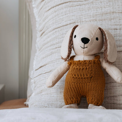 Stuffed toy dog wearing brown knitted overalls sitting on a textured surface.