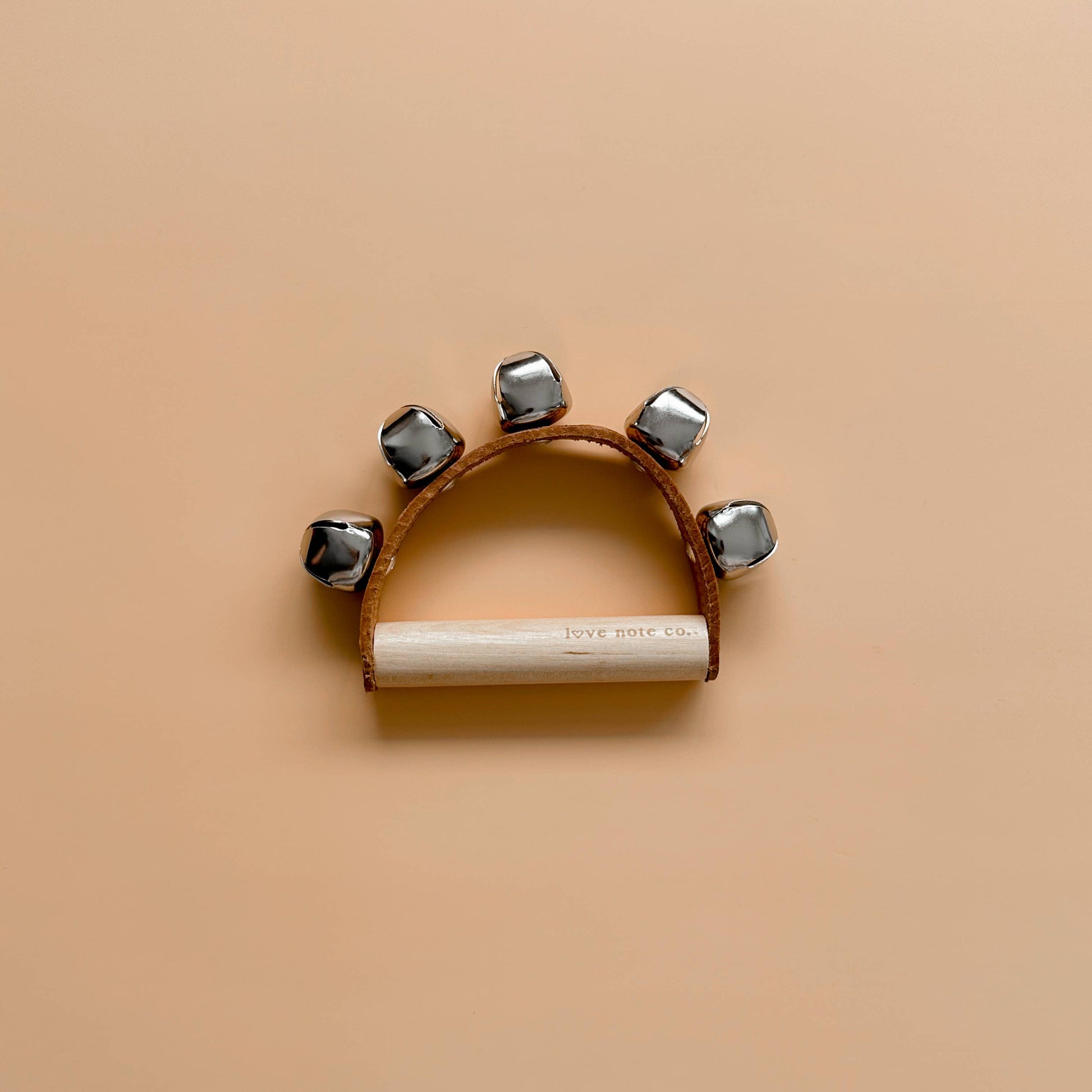 Love Note Co handbells musical toy with silver bells and a white wooden handle on a beige background.