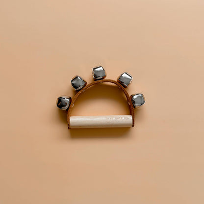 Love Note Co handbells musical toy with silver bells and a white wooden handle on a beige background.