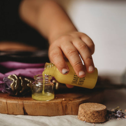 Hand pouring liquid from a yellow bottle labeled 'Wishing Well' into a small glass jar on a wooden surface with lavender and pinecones.